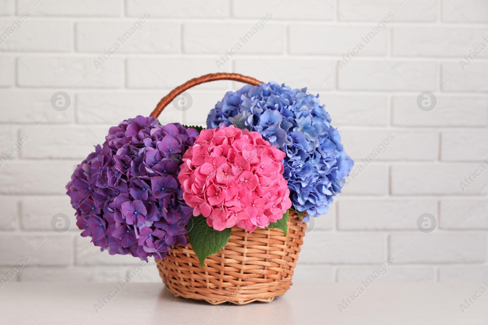 Photo of Bouquet with beautiful hortensia flowers in wicker basket on white wooden table near brick wall