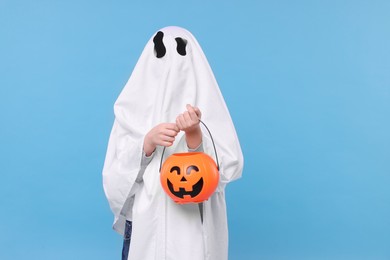 Photo of Child in white ghost costume holding pumpkin bucket on light blue background. Halloween celebration