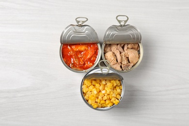 Photo of Open tin cans with sweet corn and fish meat on light table, flat lay