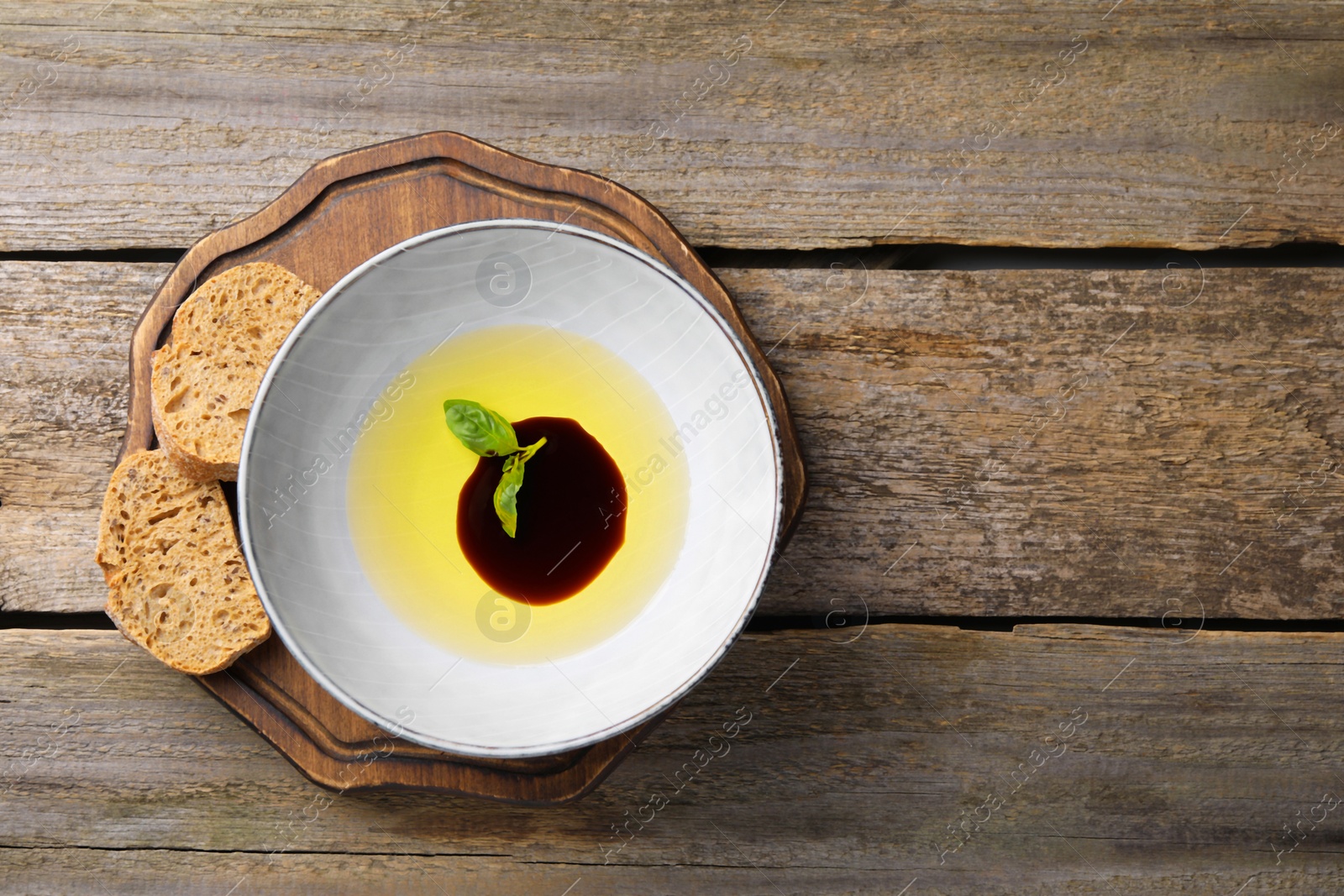 Photo of Bowl of balsamic vinegar with oil and basil served with bread slices on wooden table, top view. Space for text