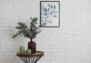 Photo of Eucalyptus branches and candles on table near white brick wall with picture. Space for text