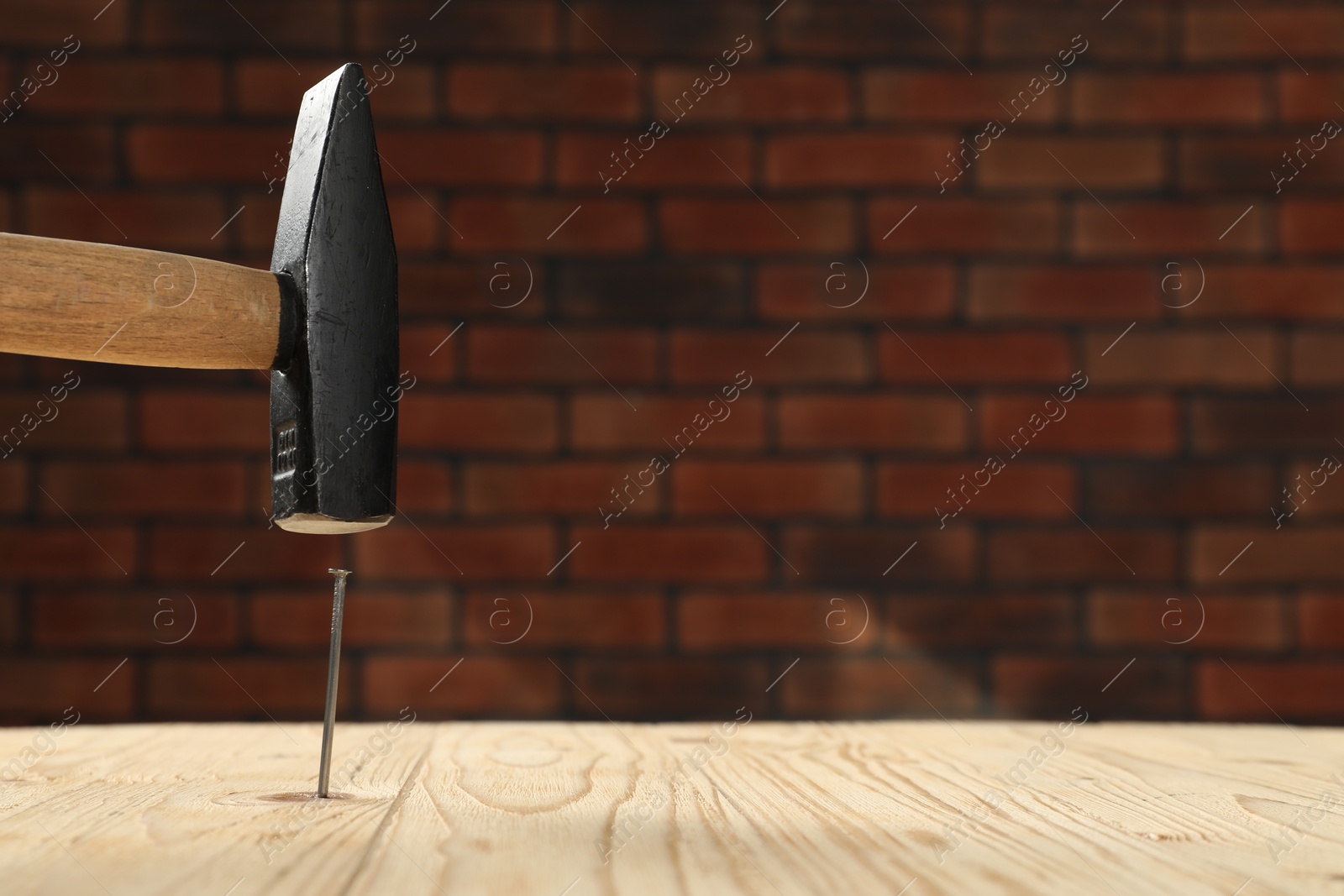 Photo of Hammering metal nail on wooden table against brick wall. Space for text