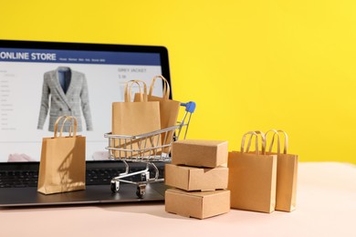 Photo of Online store. Laptop, mini shopping cart and purchases on beige table against yellow background, space for text