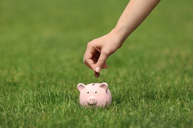 Photo of Young woman putting coin into piggy bank on green grass outdoors, closeup