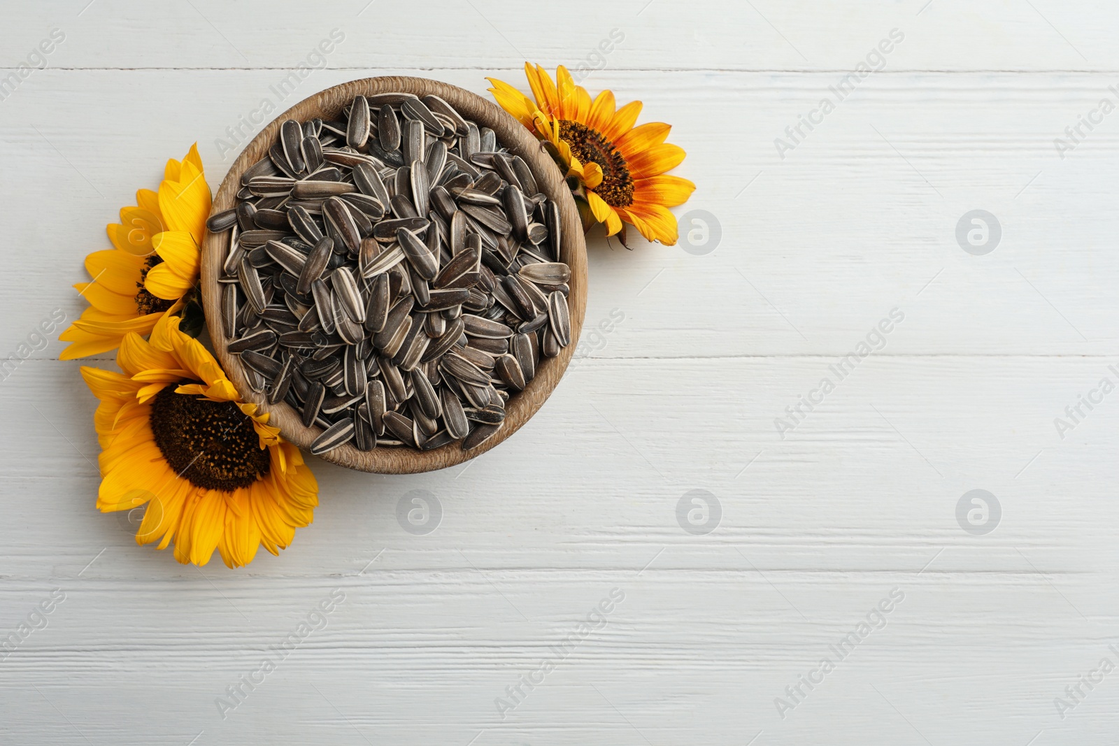 Photo of Raw sunflower seeds in bowl and flowers on white wooden table, top view. Space for text