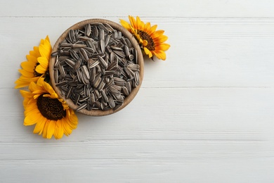 Photo of Raw sunflower seeds in bowl and flowers on white wooden table, top view. Space for text