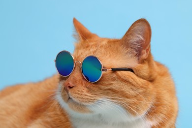 Photo of Portrait of cute ginger cat in stylish sunglasses on light blue background, closeup