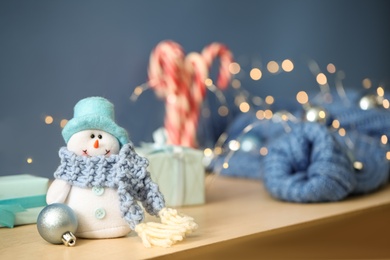 Photo of Snowman toy with knitted scarf and Christmas ball on wooden table against blurred festive lights. Space for text