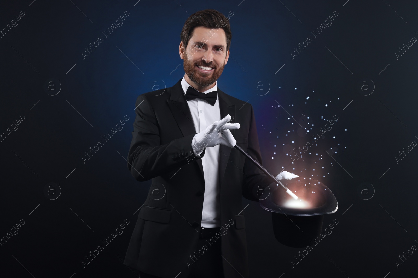 Image of Smiling magician showing trick with top hat and wand on dark blue background