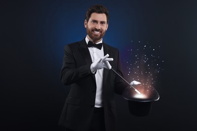 Smiling magician showing trick with top hat and wand on dark blue background