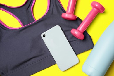 Stylish sports bra, dumbbells and smartphone on yellow background, flat lay