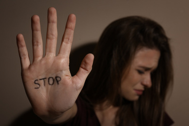 Photo of Abused young woman showing palm with word STOP near beige wall, focus on hand. Domestic violence concept