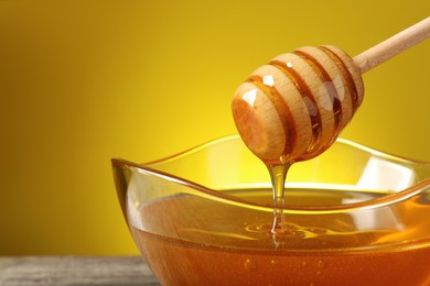 Delicious honey flowing down from dipper into bowl on table against yellow background, closeup. Space for text