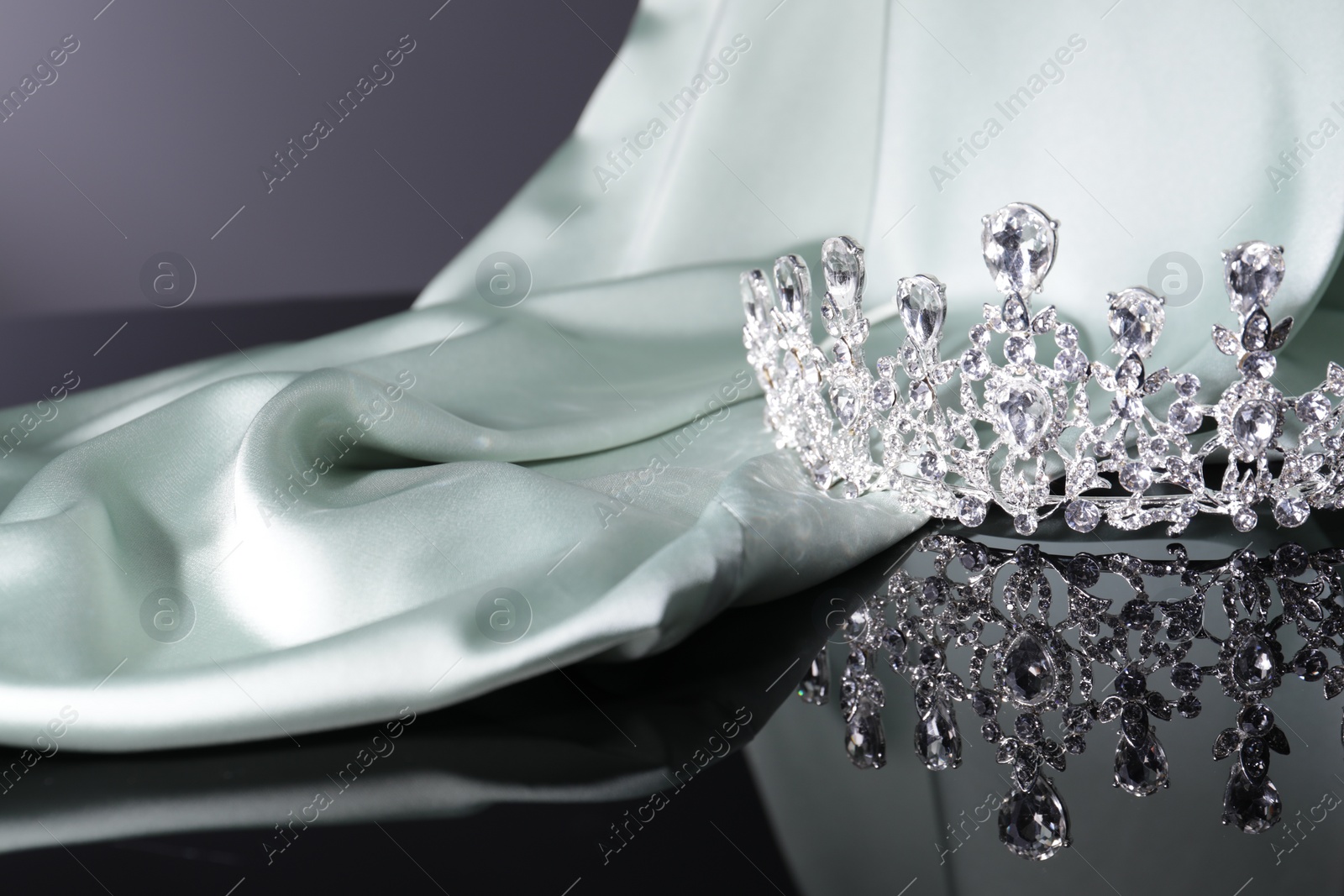 Photo of Beautiful silver tiara with gems near light cloth on dark mirror surface. Space for text