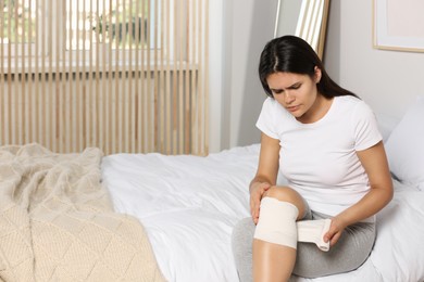 Young woman applying medical bandage onto knee in bedroom