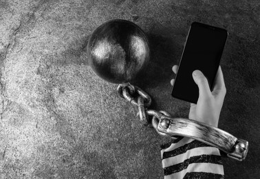 Top view of internet addicted woman in prisoner's clothes with ball and chain on her hand using smartphone at grey table, space for text. Black and white effect