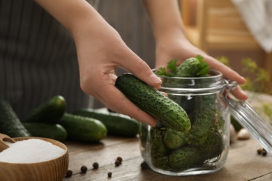 Photo of Woman putting cucumber into glass jar at wooden kitchen table, closeup. Pickling vegetables