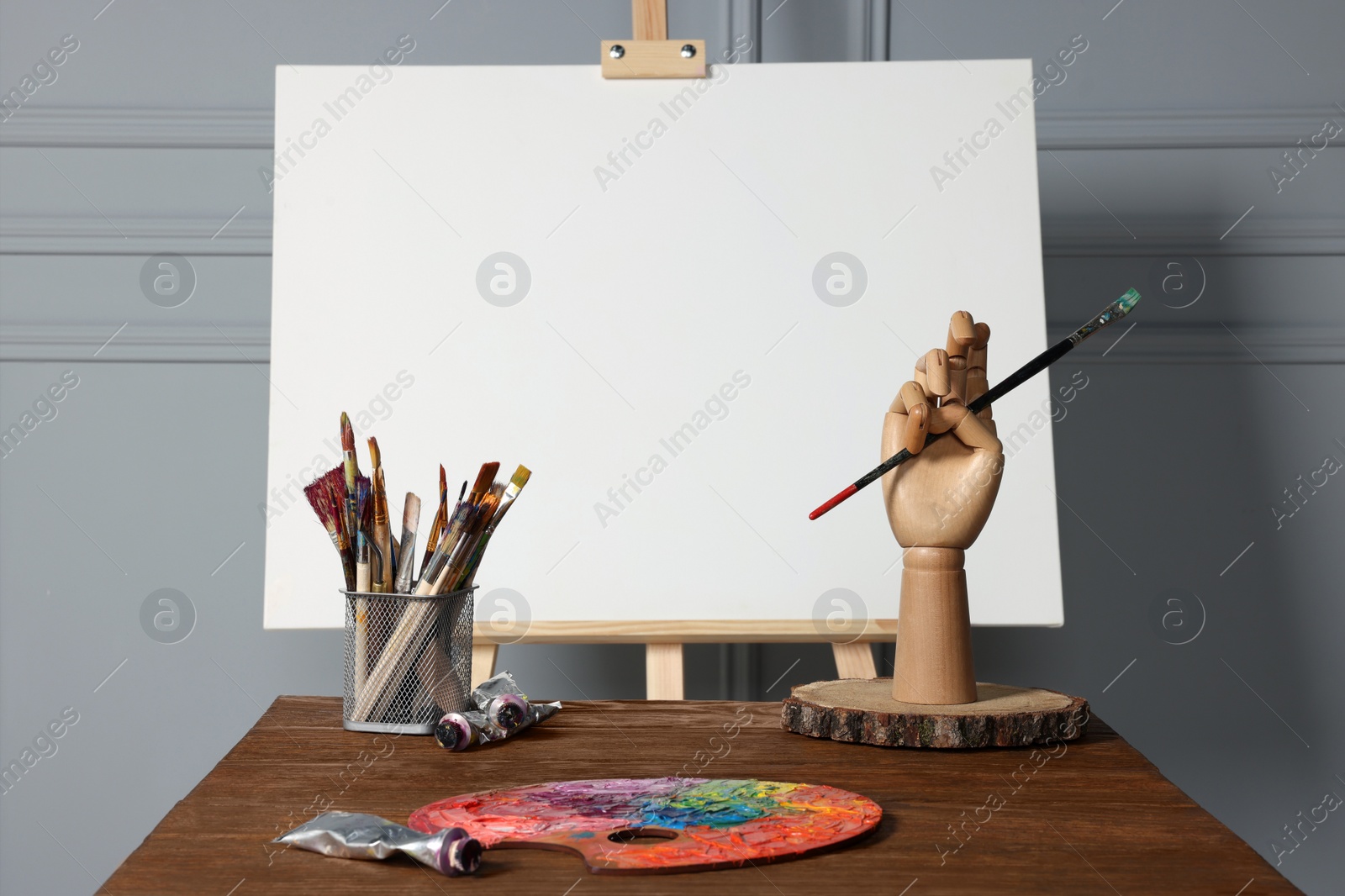 Photo of Easel with blank canvas, hand model and different art supplies on wooden table near grey wall