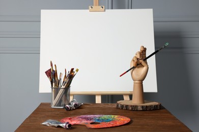 Easel with blank canvas, hand model and different art supplies on wooden table near grey wall
