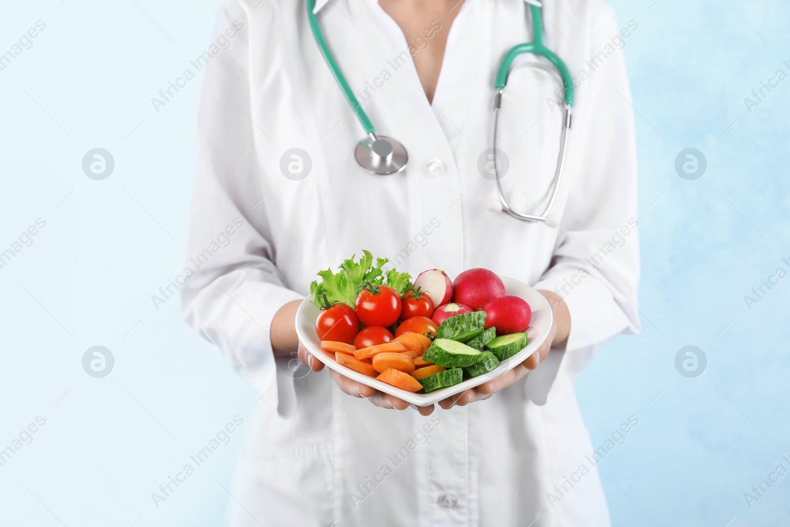 Photo of Female doctor holding plate with fresh vegetables on light background. Cardiac diet