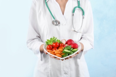 Photo of Female doctor holding plate with fresh vegetables on light background. Cardiac diet