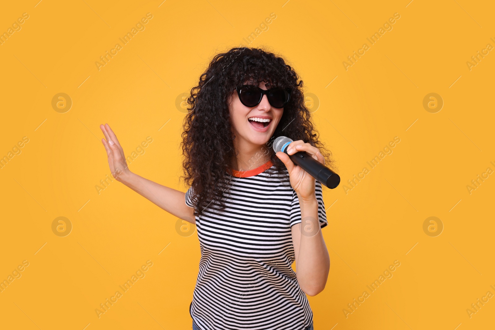 Photo of Beautiful young woman with microphone and sunglasses singing on yellow background