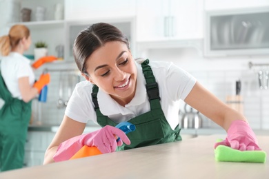 Photo of Woman using rag and sprayer for cleaning table with colleague in kitchen