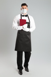 Photo of Waiter in medical face mask with menu on light grey background