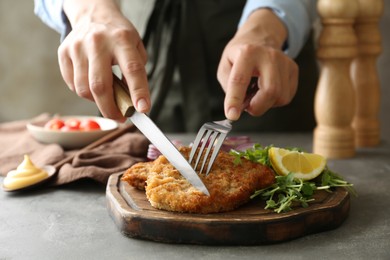Woman eating delicious schnitzel with lemon and microgreens at grey table, closeup