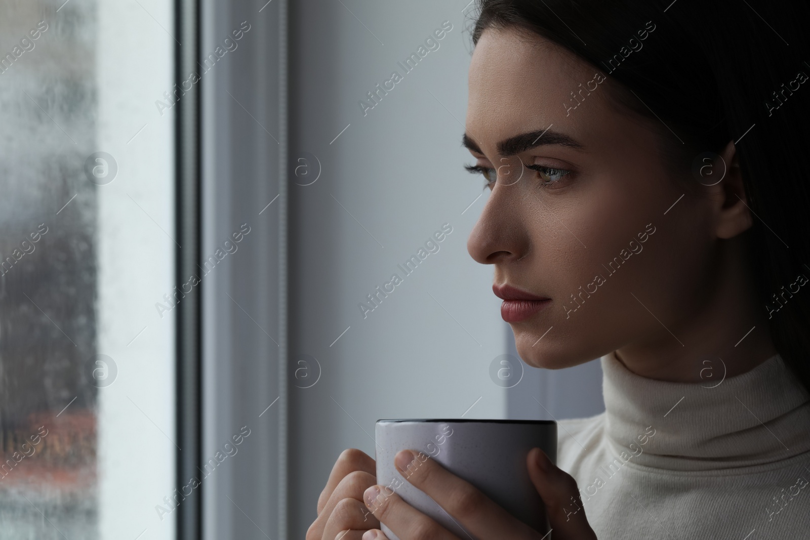 Photo of Melancholic young woman with cup of drink near window on rainy day, space for text. Loneliness concept