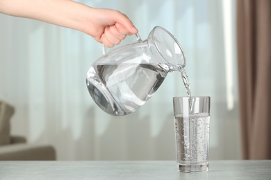 Photo of Woman pouring water from jug into glass at white table indoors, closeup