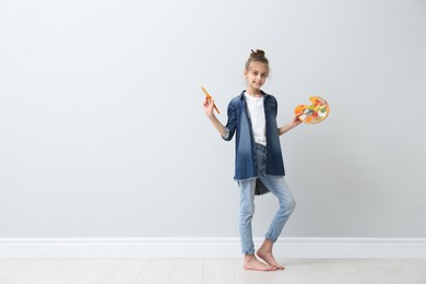 Photo of Little girl with brush and paint palette near light wall indoors. Space for text