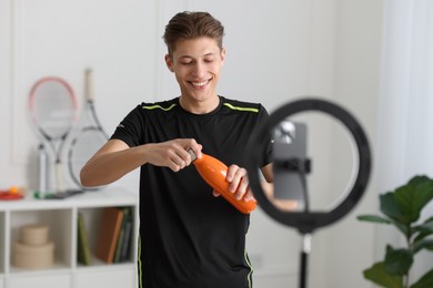 Photo of Smiling sports blogger holding bottle while streaming online fitness lesson with smartphone at home