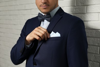 Man fixing handkerchief in breast pocket of his suit near white brick wall, closeup