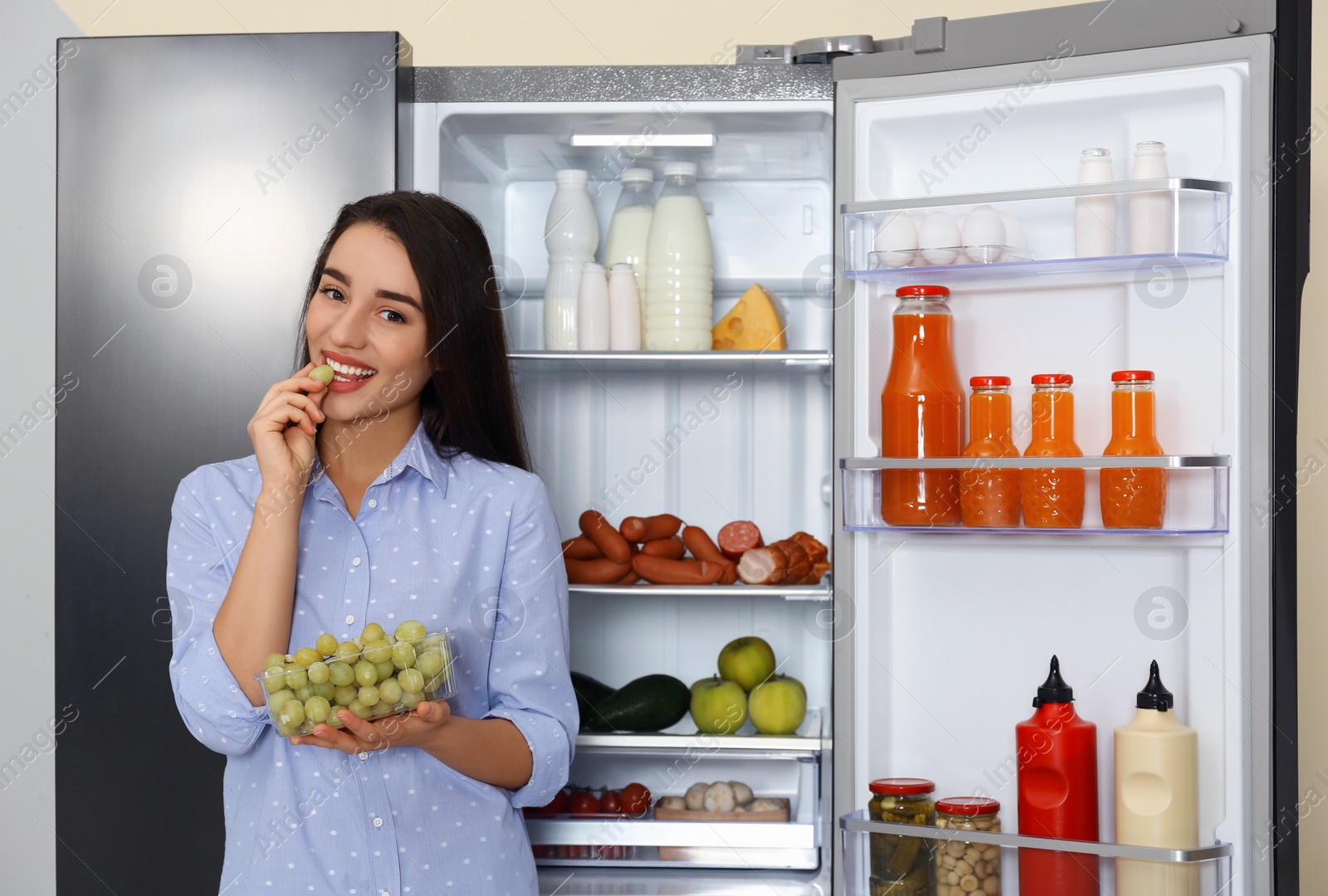 Photo of Happy young woman eating grape near open refrigerator