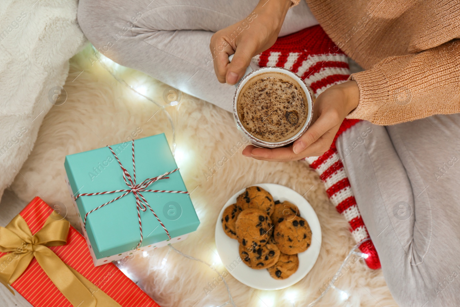 Photo of Woman with cup of hot drink, cookies and gifts on white fuzzy carpet, top view. Christmas season