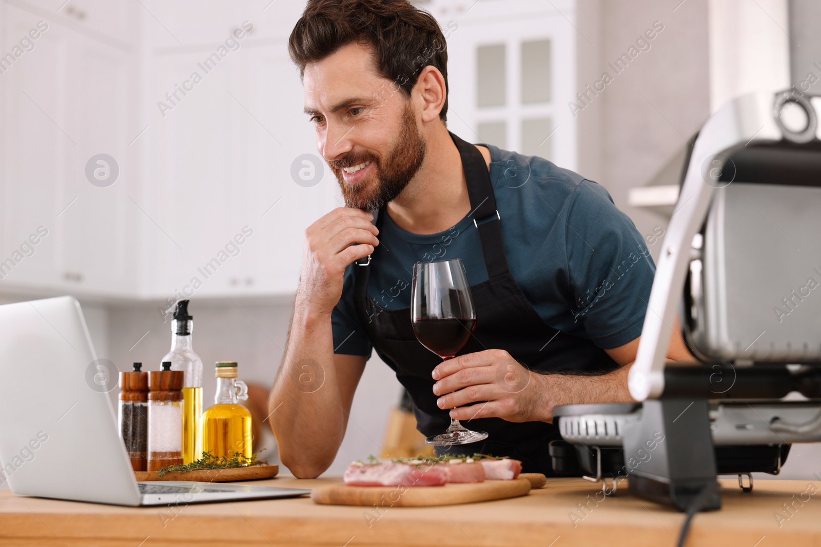 Photo of Man with glass of wine making dinner while watching online cooking course via laptop in kitchen