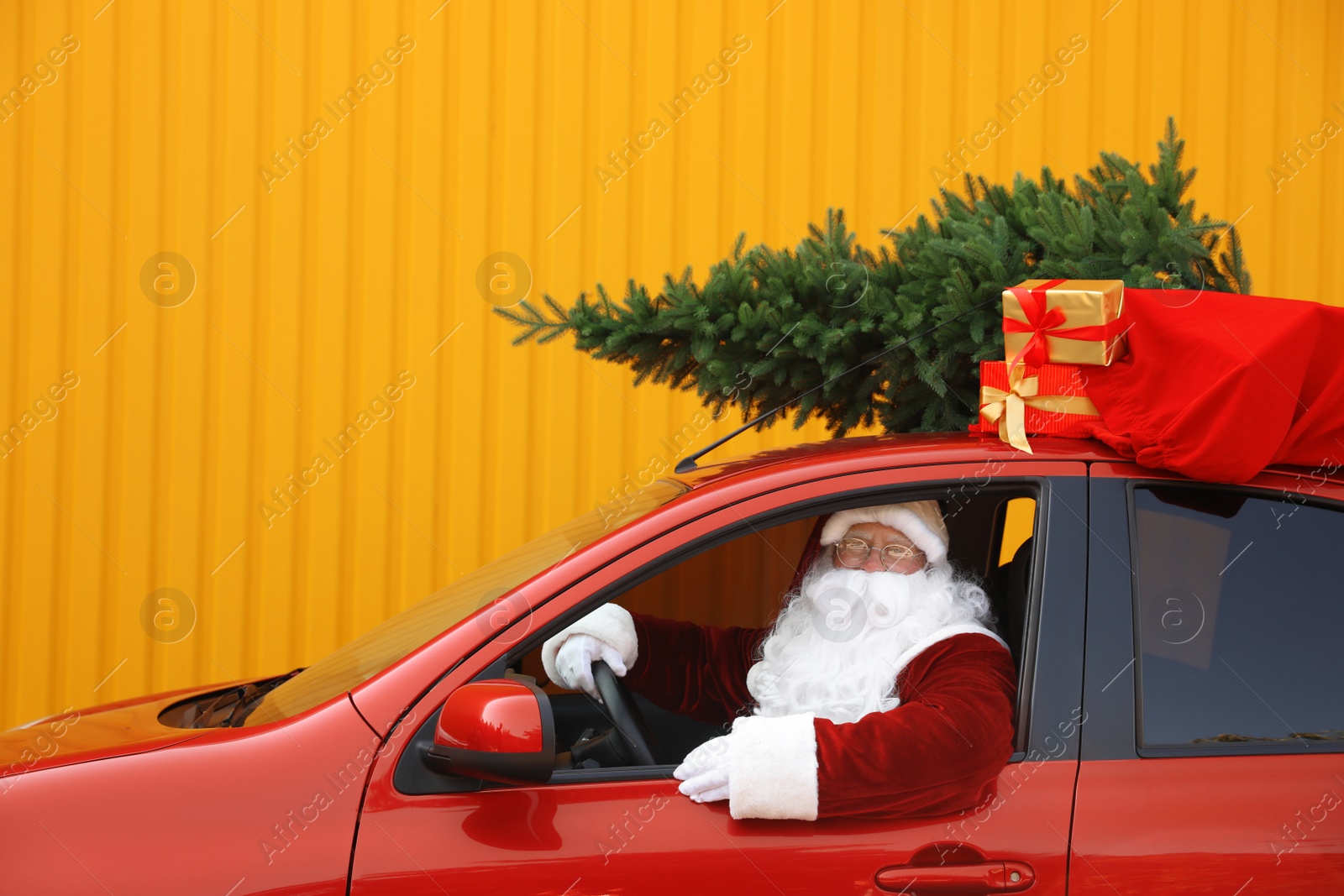 Photo of Authentic Santa Claus with fir tree and bag full of presents on roof driving car against yellow background