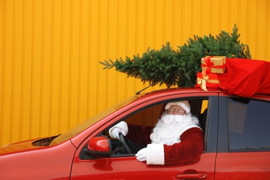 Authentic Santa Claus with fir tree and bag full of presents on roof driving car against yellow background
