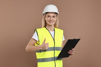 Photo of Engineer in hard hat holding clipboard and showing thumb up on brown background