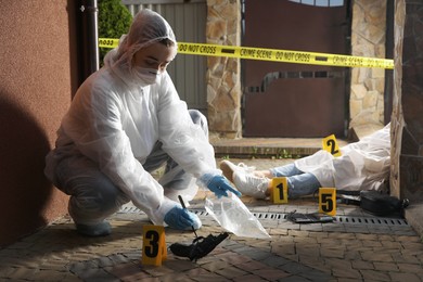 Photo of Criminologist in protective gloves working at crime scene with dead body outdoors