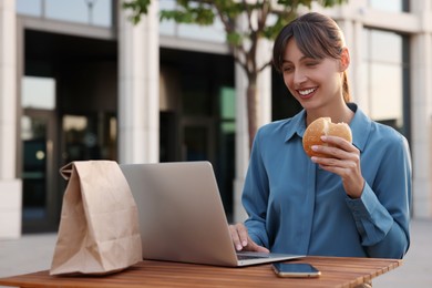 Photo of Happy businesswoman with hamburger using laptop during lunch at wooden table outdoors