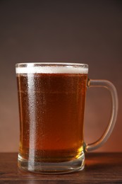 Photo of Mug with fresh beer on wooden table against brown background, closeup