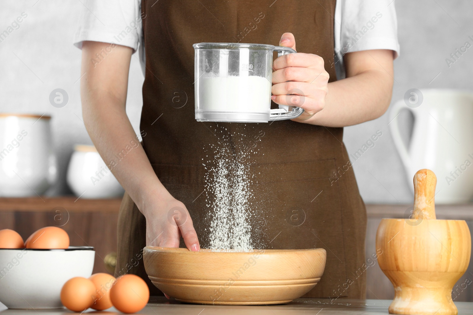 Photo of Woman sieving flour into bowl at table in kitchen, closeup