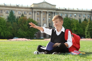 Cute boy with school stationery reading book on green lawn outdoors