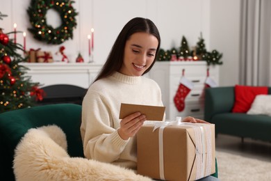 Happy young woman with Christmas gift reading greeting card at home