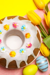 Delicious Easter cake decorated with sprinkles near beautiful tulips and painted eggs on yellow background, flat lay
