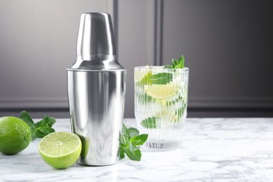 Metal cocktail shaker, delicious mojito, limes and mint on white marble table, space for text