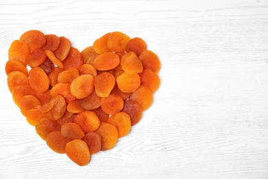 Photo of Heart shaped heap of dried apricots on white wooden table, top view with space for text. Healthy fruit