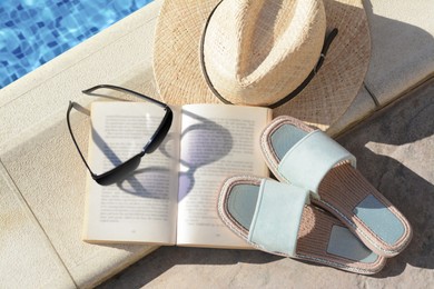 Stylish sunglasses, slippers, straw hat and book at poolside on sunny day. Beach accessories